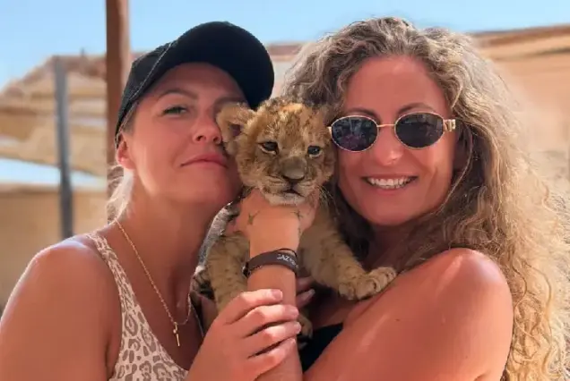 Two tourists smiling and posing closely with a cute baby lion in Hurghada